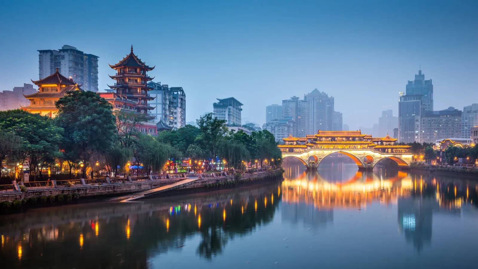 Illuminated traditional buildings and a bridge reflected in the river at dusk in Chengdu